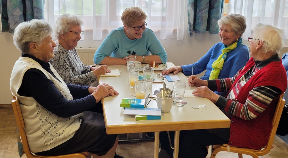 5 Personen sitzen um einen Tisch und unterhalten sich. Im Hintergrund Vorhänge.