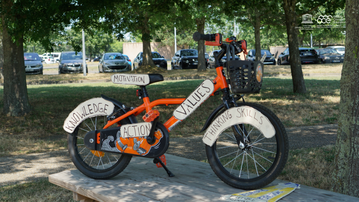 Kinder Fahrrad mit Schildern, auf denen die Ziele von Klimabildung abgebildet sind: Knowledge, Motivation, Action, Values, Thinking Skills