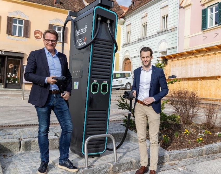 zwei Personen vor einer E-Tankstelle für KFZ, eine Person davon mit Stromzapfhahn in der Hand