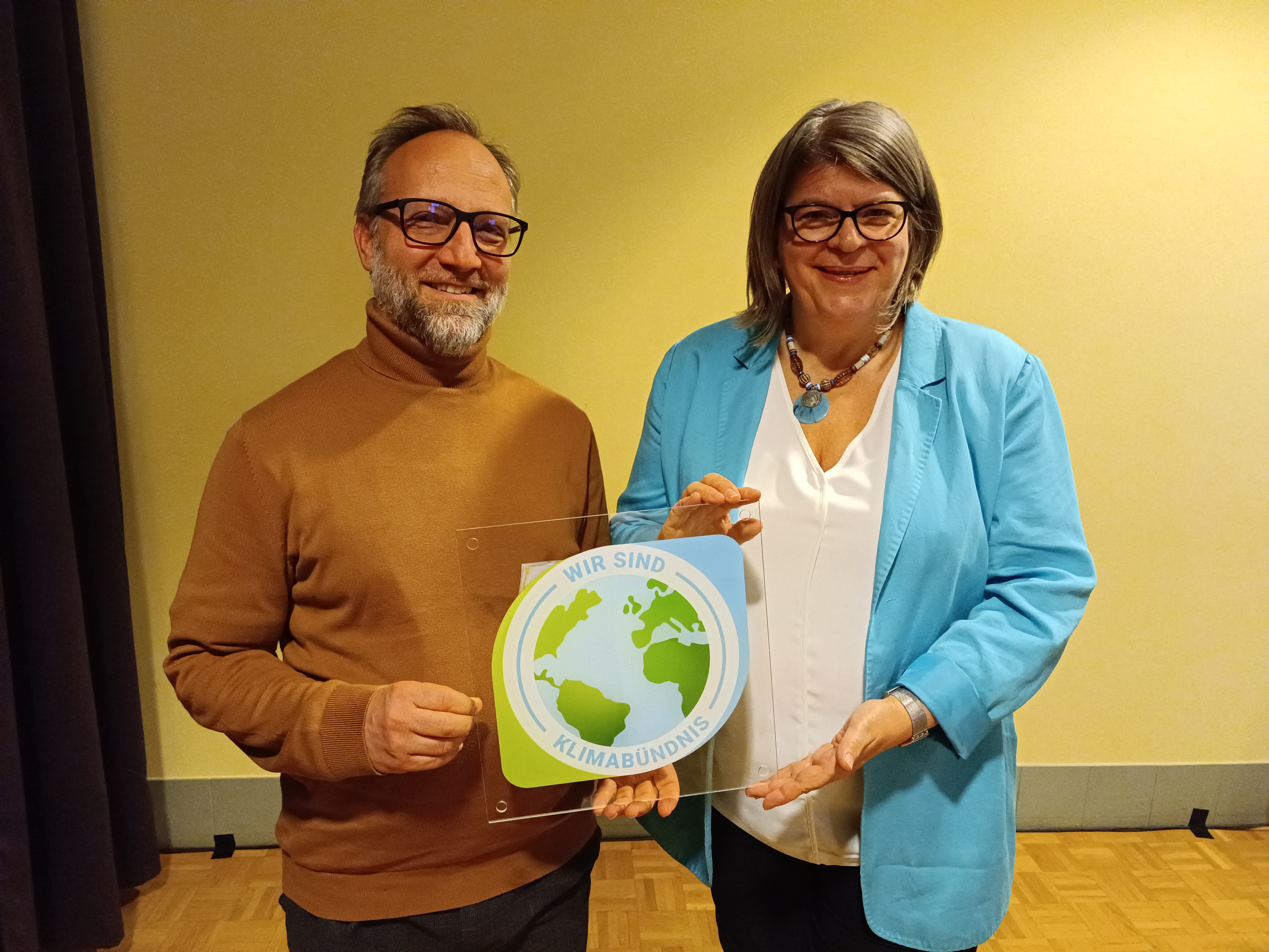Zwei Menschen stehen vor einer Wand und halten die Klimabündnis-Auszeichnung in der Hand