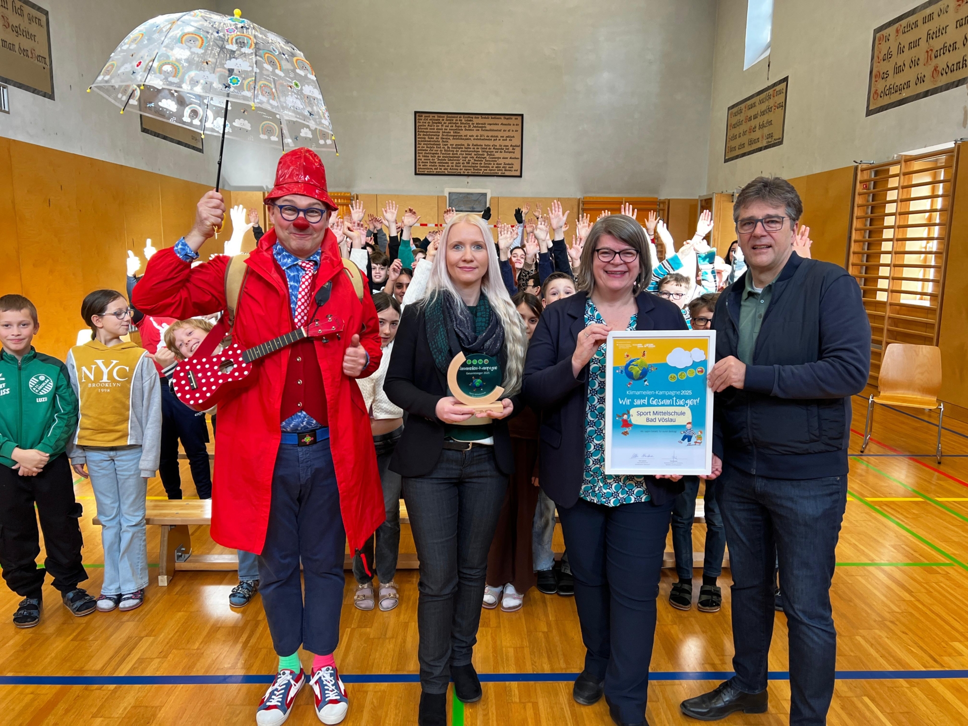 Vier Erwachsene stehen vor einer Gruppe von Kindern in einer Turnhalle und präsentieren eine Auszeichnung; der Mann ganz links trägt ein Clown Kostüm