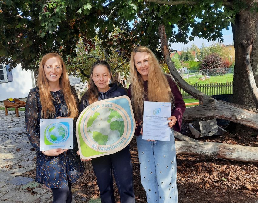 Drei Frauen stehen nebeneinander und halten zwei Urkunden und ein großes Holzschild mit dem Klimabündnis-Logo