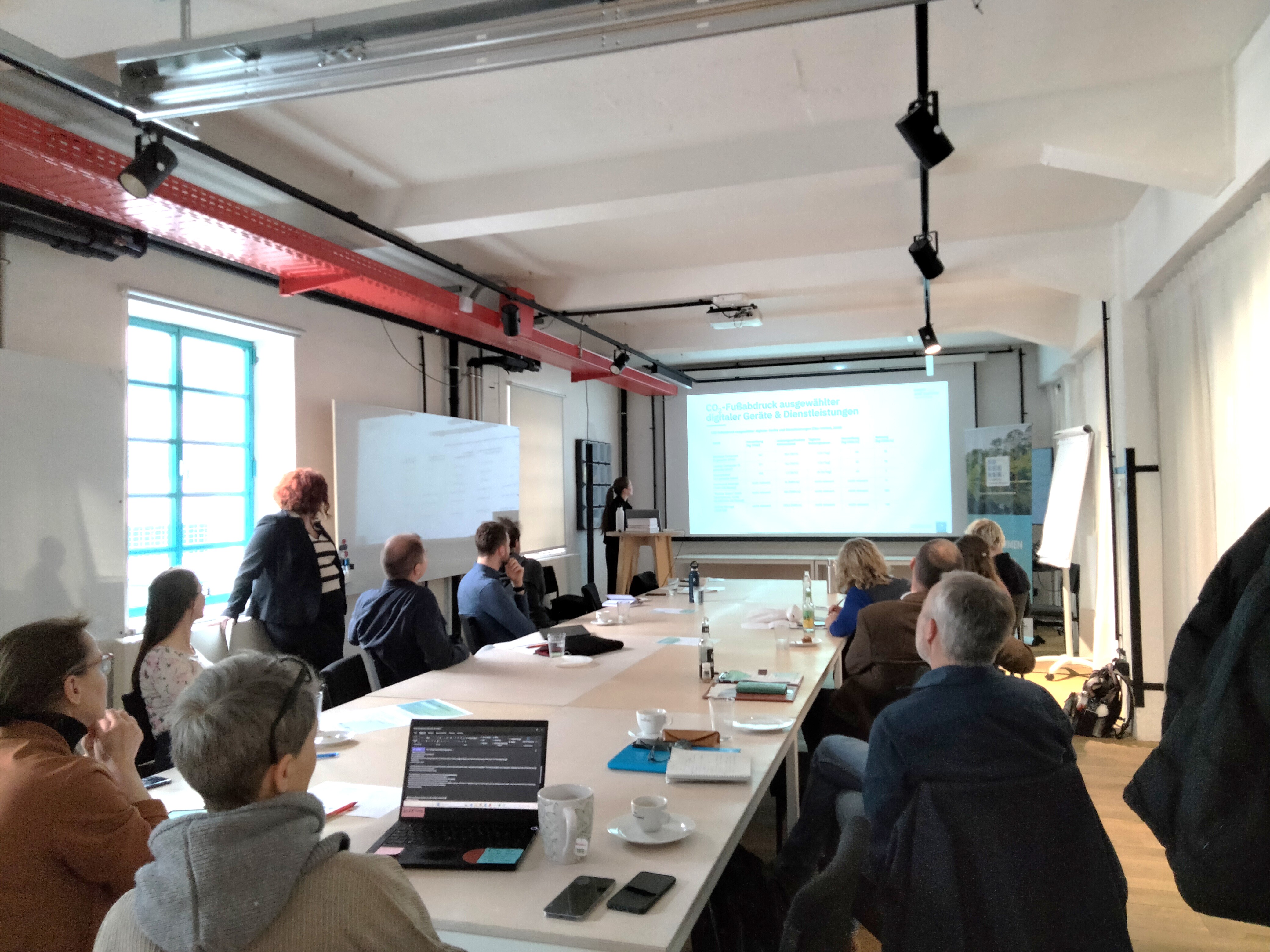 ungefähr fünfzehn Menschen sitzen um einen Konferenztisch und Blicken auf eine Präsentation